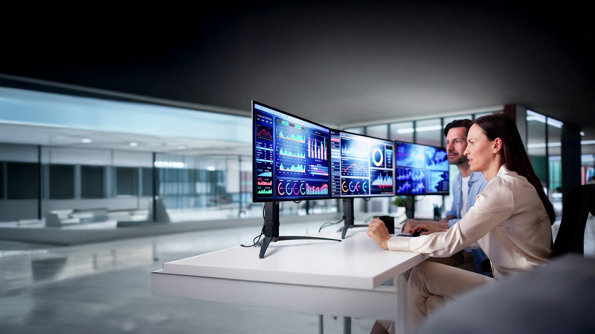 Woman and man sitting at a desk in an office, viewing three computer monitors.