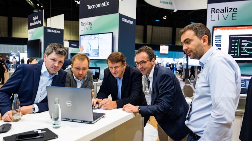 A group of businessmen in suits gather around a laptop, viewing a presentation at the Siemens Realize Live conference.