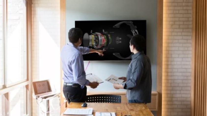 Two people standing at the front of the room while pointing at a television screen with papers in hand.