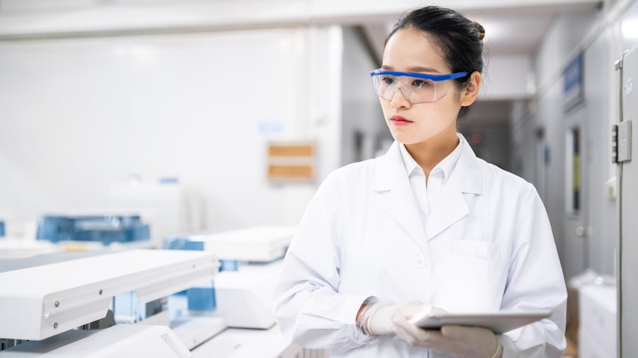 Engineer in a lab coat inspecting ongoing product testing while checking her notes on a clipboard.