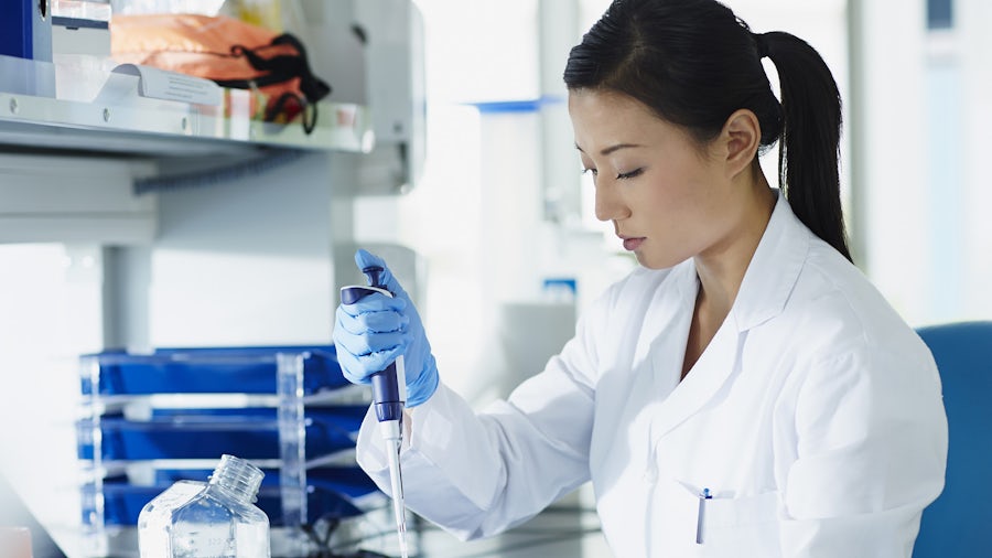 A woman working in a laboratory using a pipette to transfer liquid into a container, with scientific equipment visible in the background.