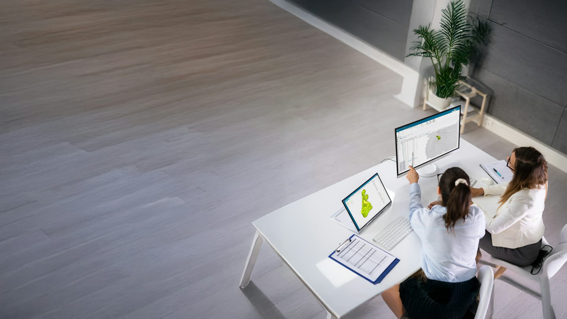 Two female coworkers collaborating at a desk with a computer monitor and tablet displaying Siemens NX CAD software.