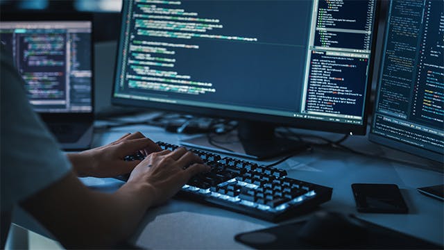 Close-up focus on a person's hands typing on a desktop computer keyboard and the screens show coding language user interface.