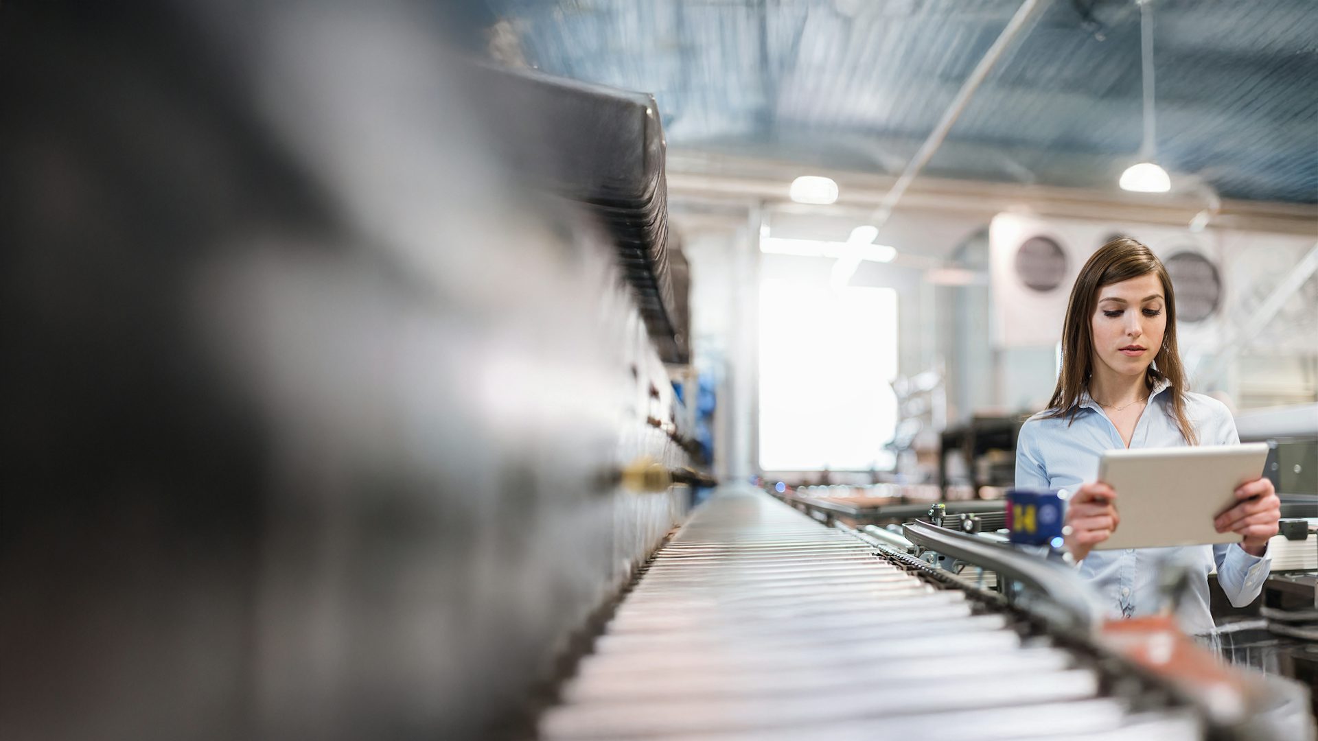 Woman looking at a laptop next to an assembly line in a factory, representing cloud-based Quality Management System