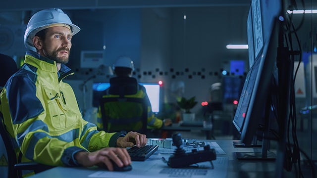 A man dressed in a yellow construction jacket and a white hard hat is sitting in front of computer monitors in a dark office setting.