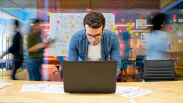 Man working on a laptop in an office, with a large whiteboard covered in sticky notes and people moving in the background.