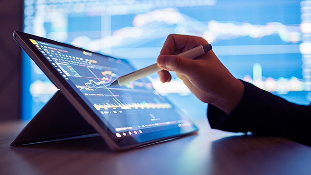 Hand using a stylus on a tablet with a large display screen in the background.