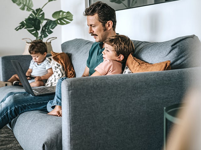 Man sitting on couch working on a laptop, with two children sitting nearby