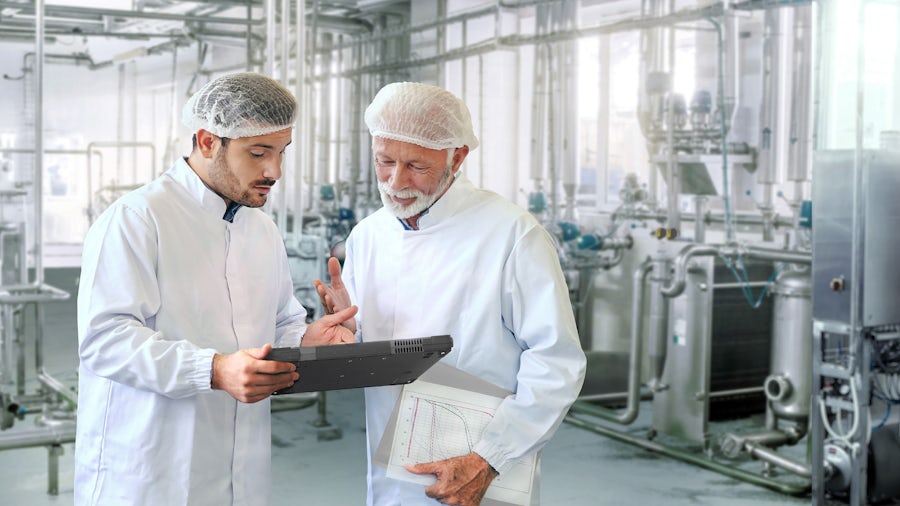 Two men in hairnets and white coats look at a tablet while standing in a food packing facility.