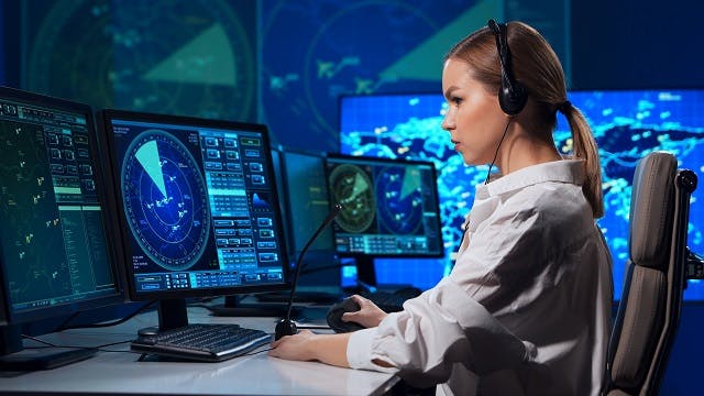 A woman wearing headphones sits at a desk monitoring an aerospace communications network on an array of digital displays.