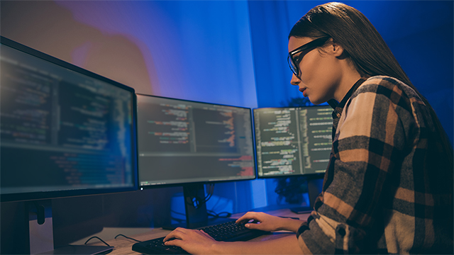 Female programmer intently reviewing code on multiple monitors at her desk.