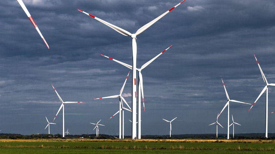 More than a dozen white wind turbines standing in an open field on a dark gray cloudy sky.