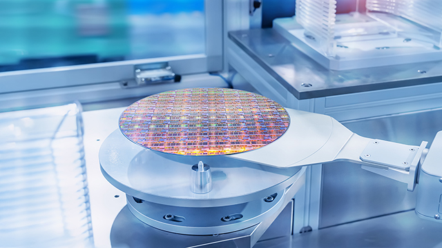 Silicon wafer with colorful iridescent surface mounted on white testing platform. The wafer is surrounded by precision mechanical equipment in a clean room environment with blue-tinted lighting.