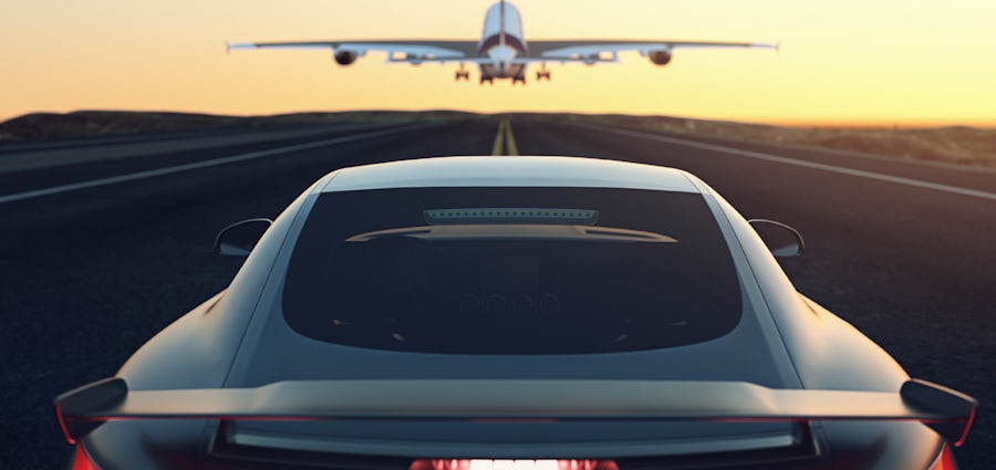 A car drives on the road while an airplane flies overhead in a clear blue sky.