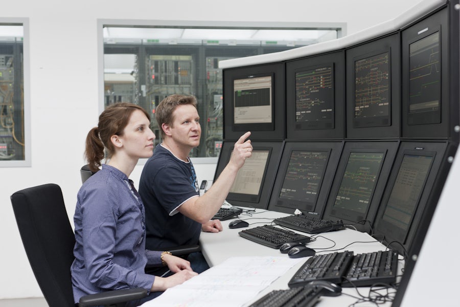 a man and a women sit at a desk surrounded by many computer screens where they are looking at, discussing, and possibly analyzing data at a logistics center