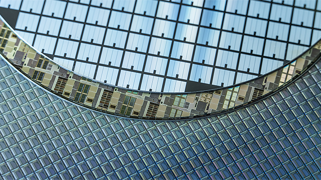 Close-up of a circular silicon wafer with a complex grid of microchips and circuits, showing intricate geometric patterns and lines. The wafer has a reflective, metallic surface with blue and green tones, highlighting the fine details of semiconductor technology.