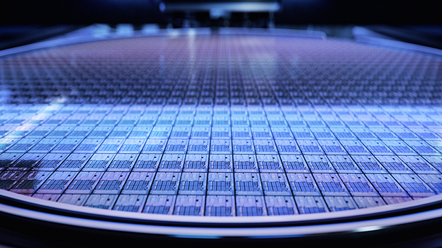 A silicon wafer with blue-tinted lighting, showing detailed grid pattern of integrated circuits across its surface. The perspective emphasizes the wafer's circular shape and precise geometric patterns.