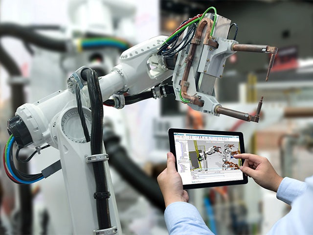 Ingenieur mit Tablet, das ein 3D-Robotik-Simulationsmodell vor realem Roboter in der Fabrikhalle anzeigt.