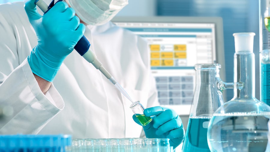 Research worker performing development activities using liquid chemicals in a laboratory setting. RD&L software appears on a computer screen in the background.