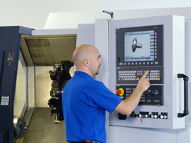A man in a blue polo clicking a button on a manufacturing machine. The screen is showing a part to be manufactured.