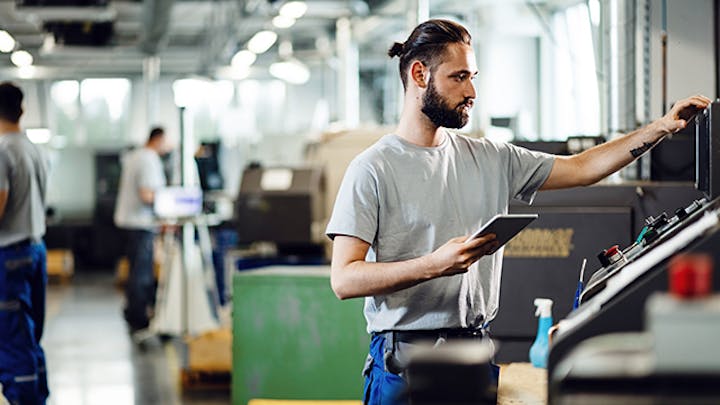 Image of a man in a white shirt working in a factory. He is holding a tablet and examining a piece of equipment. This image is intended to convey the concept of nonconformance management.