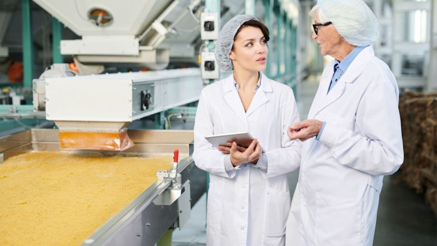 workers in lab coats discuss recipe execution next to industrial food processing equipment