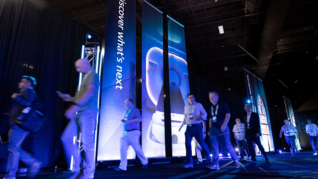 Solutions Center at Realize LIVE Americas. Attendees walking through a hallway with banners in the background that are lit up. 
