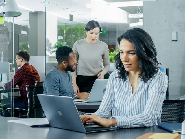 people working in a workspace on their laptops where there are two that have a discussion. 