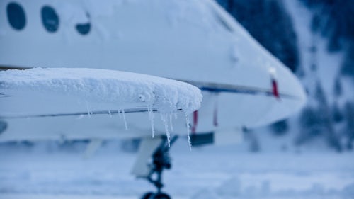 The front of a passenger plane in need of aircraft de-icing is shown in a snowy setting, with frost and icicles encrusting the tip of the wing