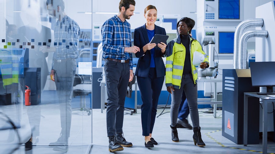 Three engineers walk across a manufacturing floor. The middle engineer holds a tablet.