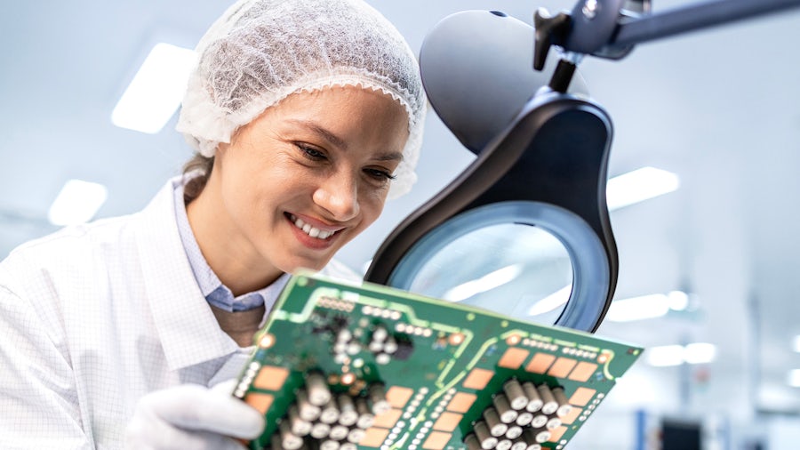 A lab technician looks over a magnifying glass at a large electronics component with chips.