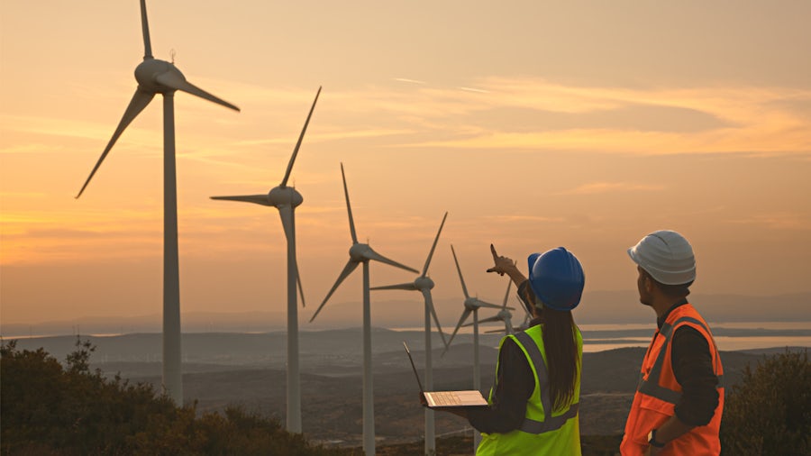 A man and woman in high visibility vests and hard hats look at a row of wind turbines standing on a ridge overlooking a riverbed and forest.