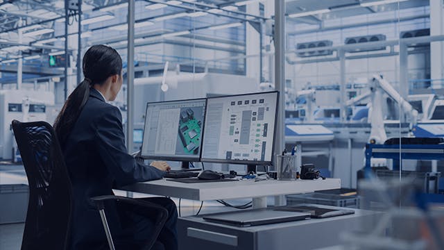 Female engineer working at a desk with two monitors in a robotic arm factory.