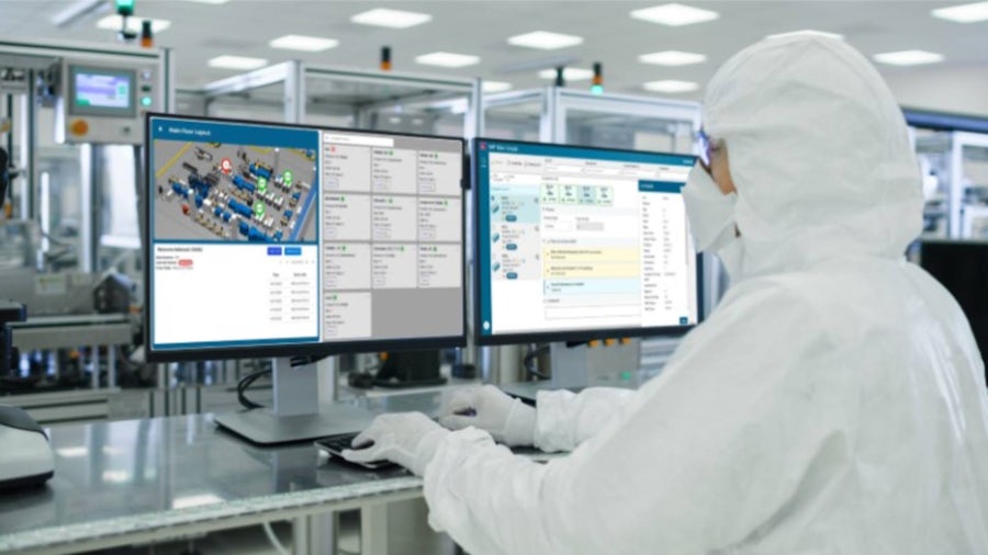 A worker monitors manufacturing processes through multiple computer displays. The screens show production monitoring software with layout views and control interfaces, while manufacturing equipment is visible.