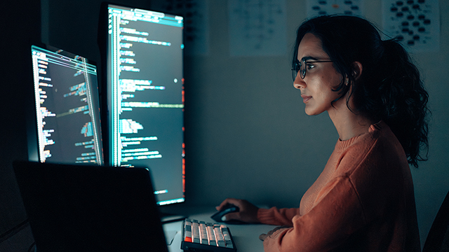 Female programmer coding at night in a high-tech workspace with a laptop and multiple monitors.
