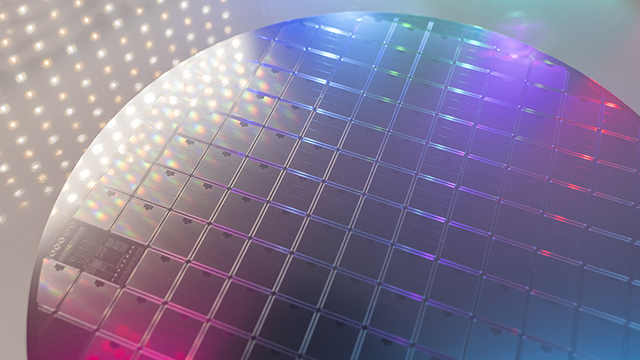 Silicon wafer under specialized lighting showing a precise grid pattern with holographic diffraction effects. White LED array visible in background creates a gradient of colors from white to blue and pink across the wafer surface.