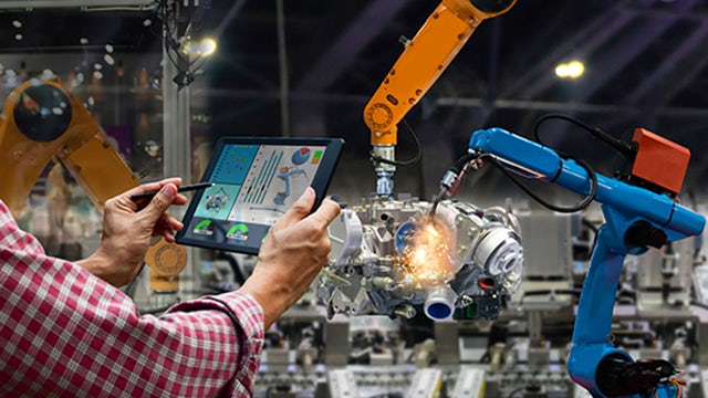 A man uses a tablet to control two robotic arms on the floor of a manufacturing facility