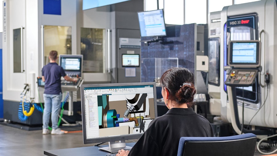 Two workers are shown on the shop floor, one in the foreground on a computer looking at parts in a software program, the other in the background monitoring a machine