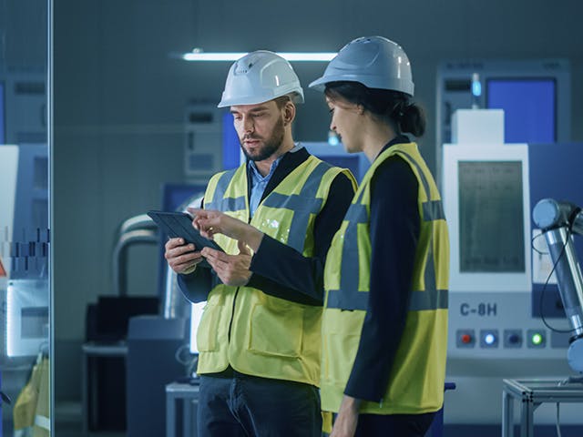 A man in a hard hat holds a tablet while a woman in a hard hat points to the tablet screen