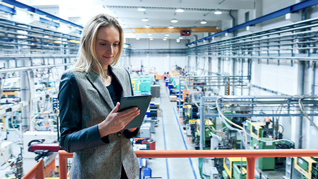 A woman looks at a tablet computer, with a factory floor behind her