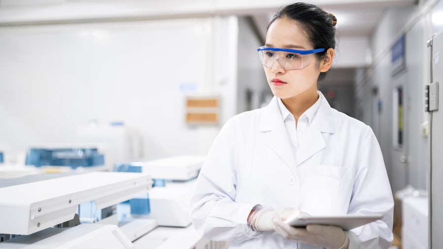 A scientist wearing a lab coat and protective eyewear while holding a tablet and walking through a lab.