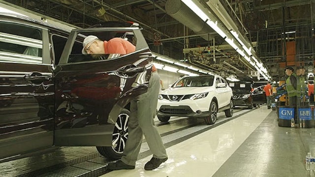 A vehicle manufacturing worker opening the door to a black SUV on the line, with a white SUV and another black SUV on the line.