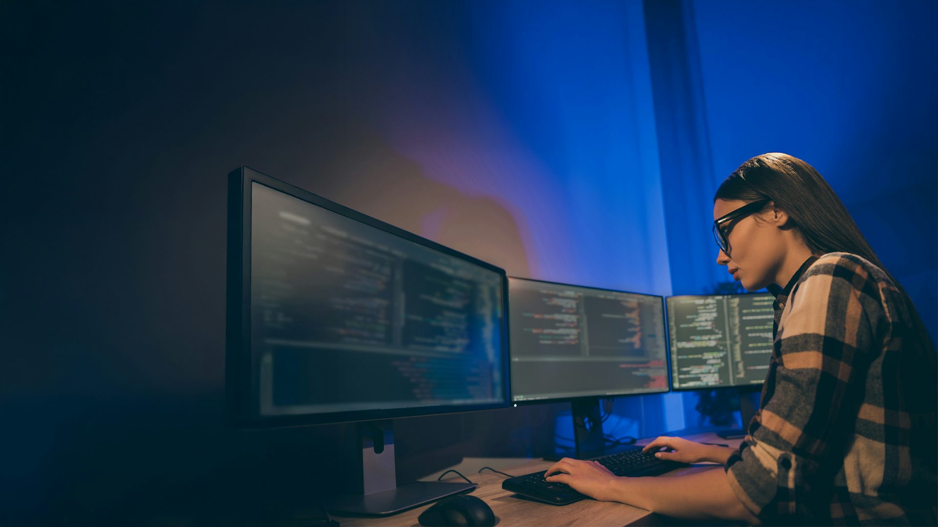 Female programmer intently reviewing code on multiple monitors at her desk.
