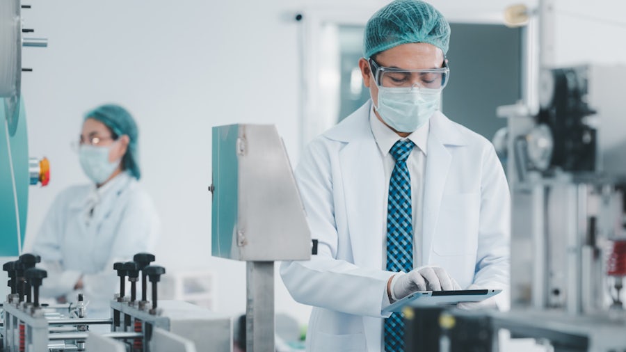 Man working on a tablet in a medical device manufacturing environment with a second female worker in the background. 