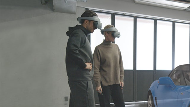Un hombre y una mujer, cada uno con un casco de realidad virtual de Sony, utiliza el software Siemens NX Immersive Designer para trabajar en un modelo virtual de un automóvil deportivo azul.