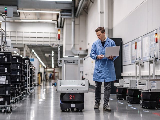 A man in a lab coat holding a laptop walks next to an autonomous robot on a factory floor