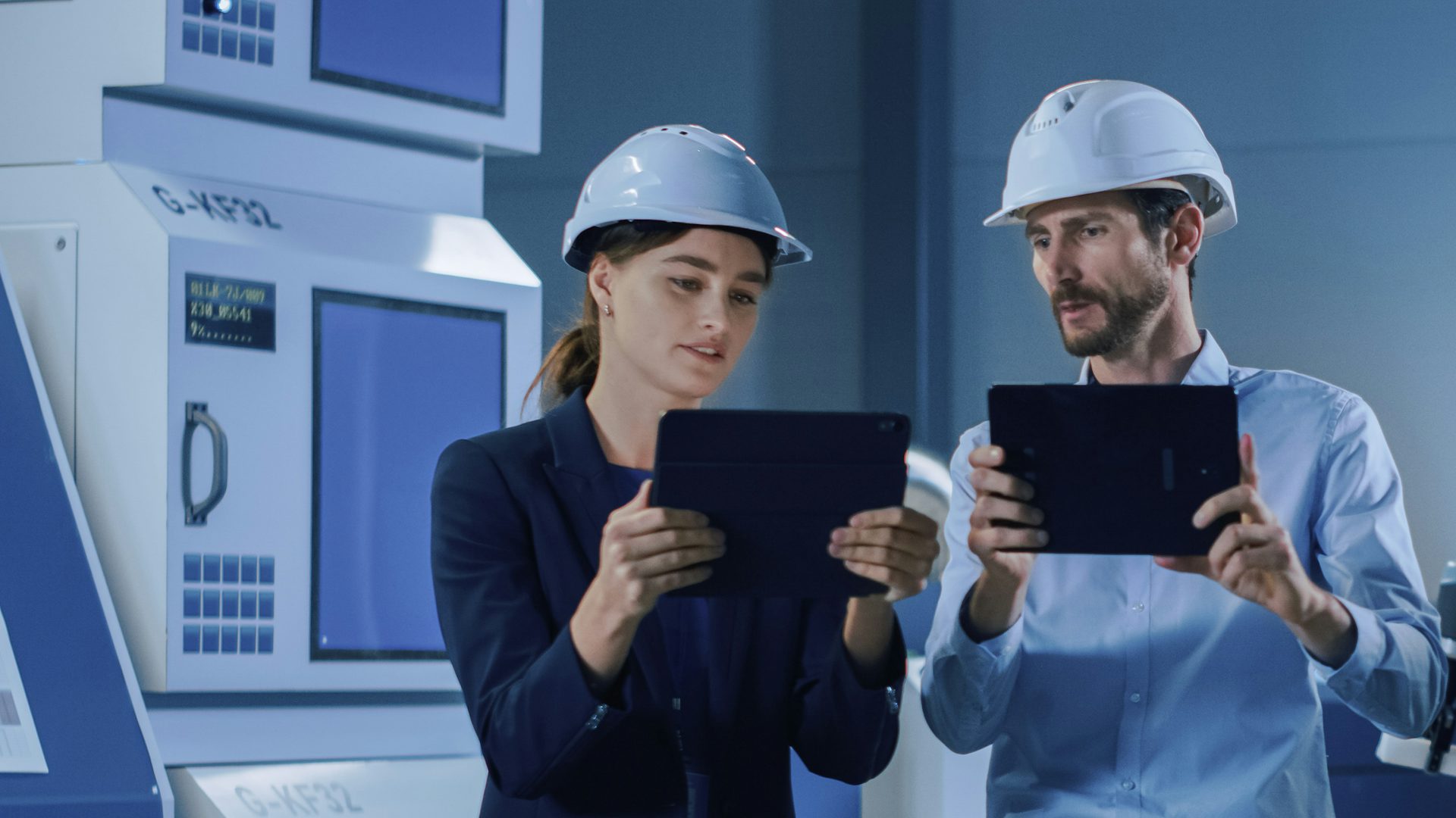A man and a woman in hard hats, each holding a tablet, on a factory floor