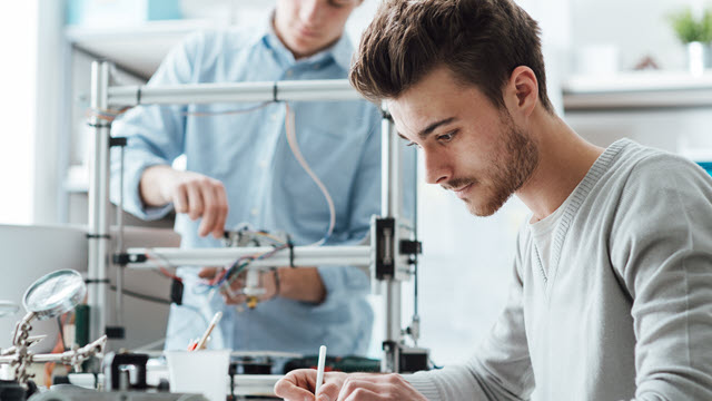 Engineer is sitting on desk and writing, while other one is standing and working on hardware device