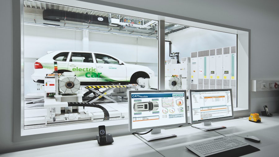 A vehicle's durability being tested in a lab with a computer desk set up in another room, looking at the test through windows. 