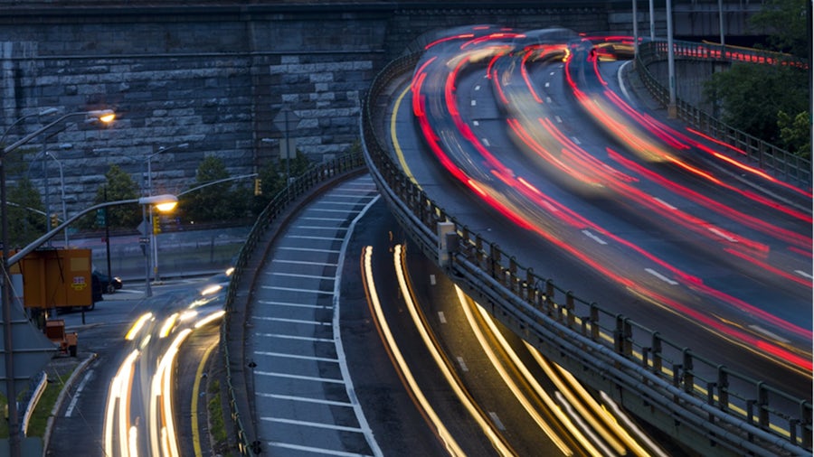 Long-exposure photo of a multi-lane highway at dusk, showing bright red and white streaks of light from moving cars, highlighting the motion and speed of traffic on a curved, elevated road against a stone wall background.